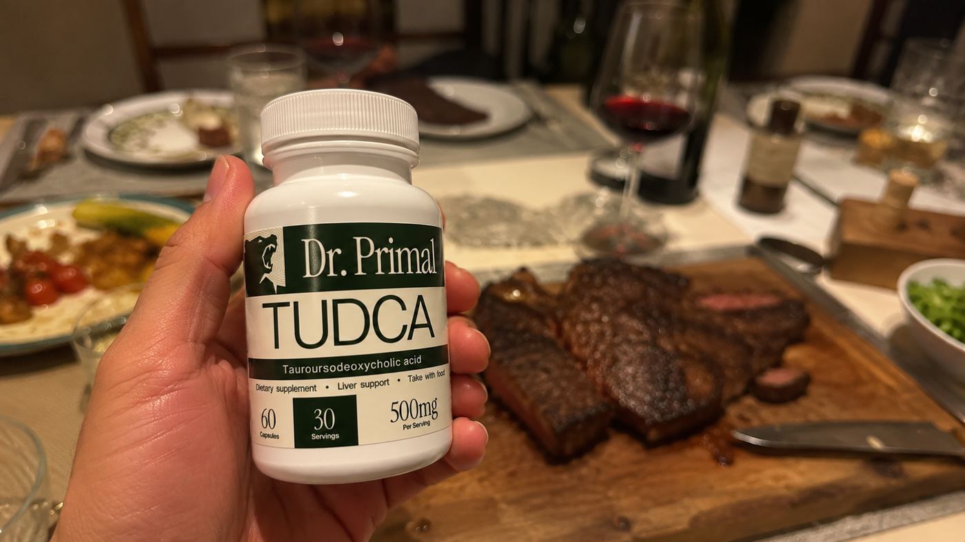 Holding a jar of Dr. Primal TUDCA capsules over a dinner table with steak